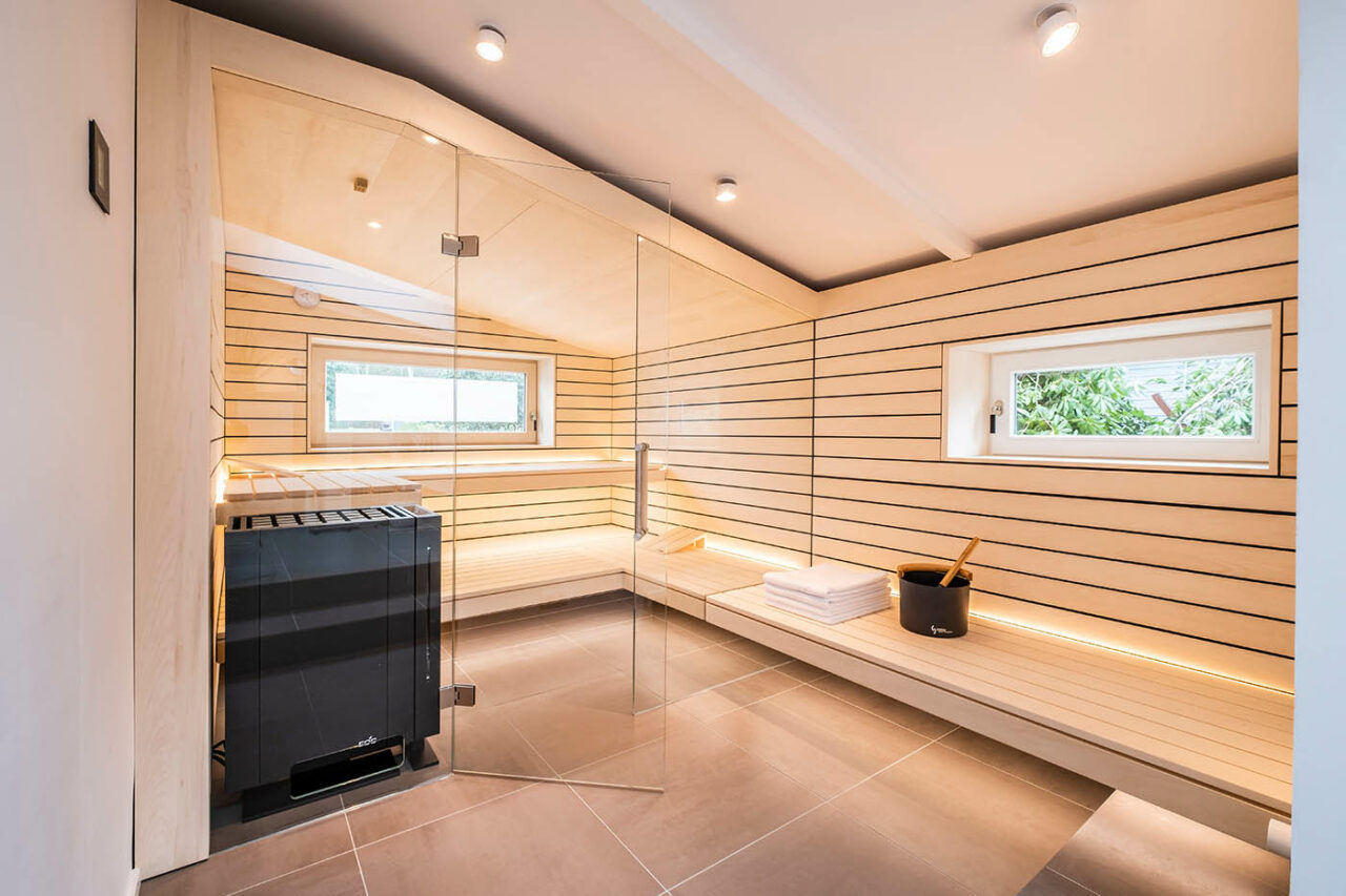 Saunabau Hannover | Nr. 1 für Sauna nach Maß 11 Design Sauna mit Fenster nach draußen. Sauna mit Dachschräge nach Maß in hellem Holz. Große Glasfront, nach außen verlängerte Bänke und LED Beleuchtung. Frei stehender Saunaofen und schwebende Saunabänke. Saunabau Hannover. Saunabau Niedersachsen.