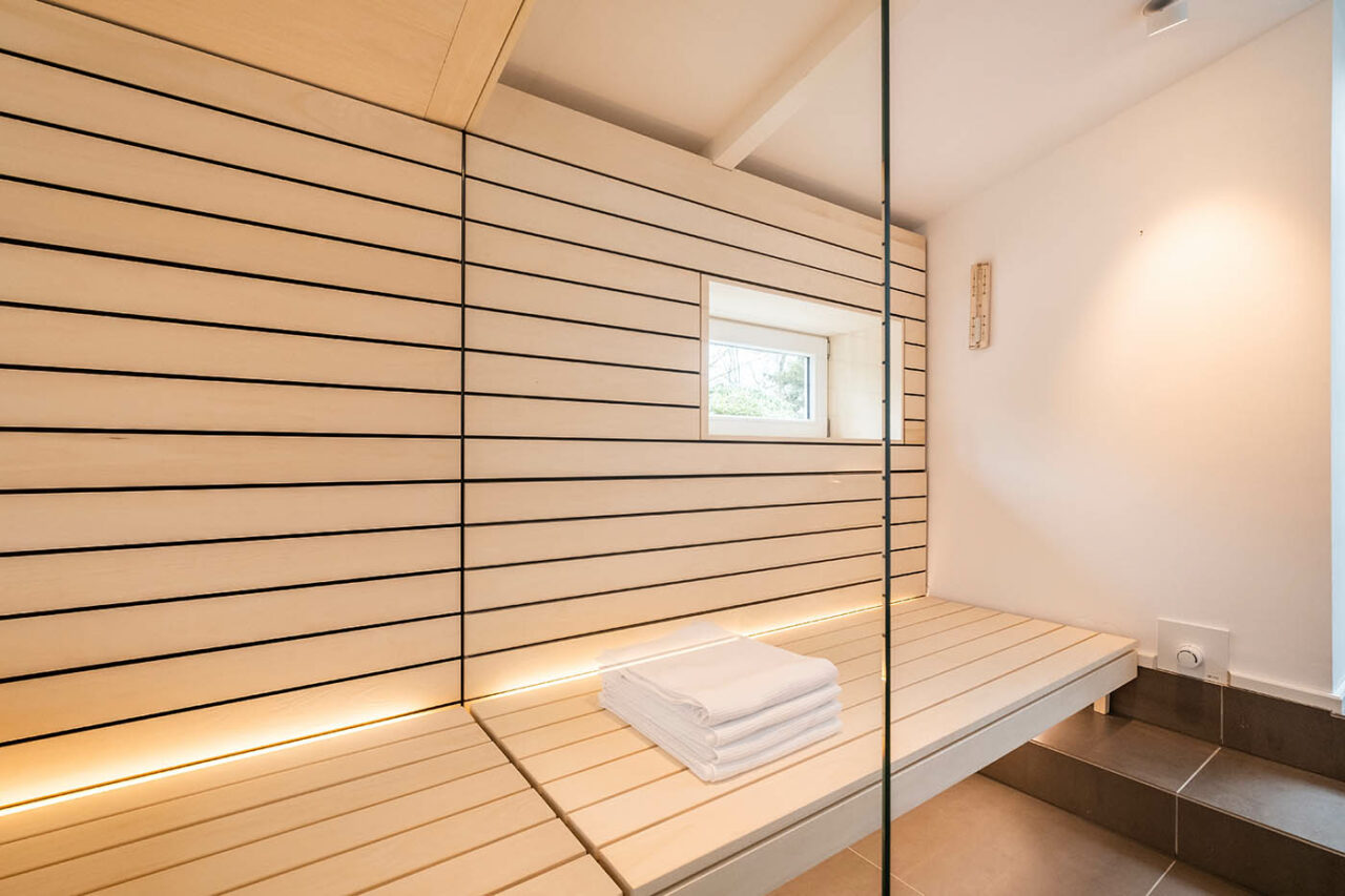Sauna mit Fenster nach draußen 5 Design Sauna mit Fenster nach draußen. Sauna mit Dachschräge nach Maß in hellem Holz. Große Glasfront, nach außen verlängerte Bänke und LED Beleuchtung. Frei stehender Saunaofen und schwebende Saunabänke.