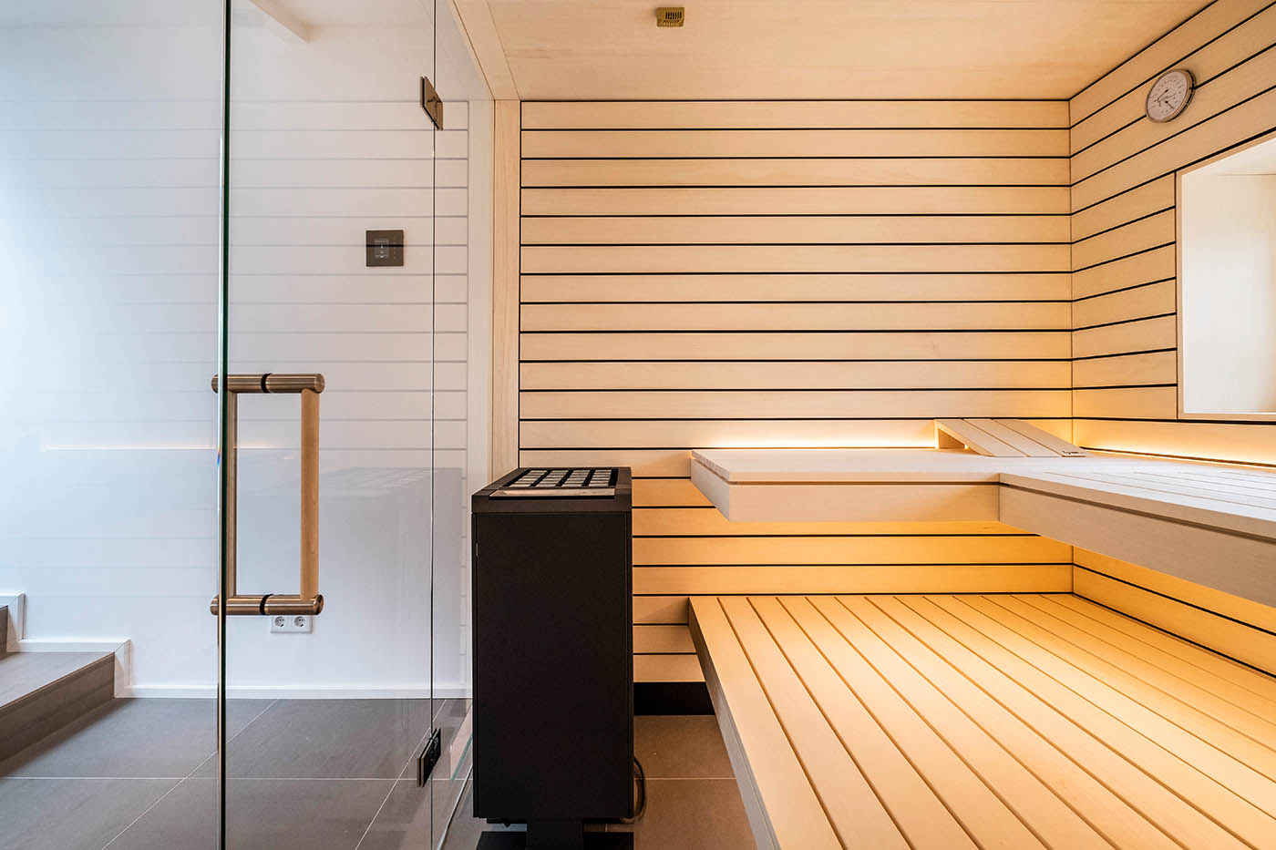 Saunaholz: Espe Doppelnut | schwarze Feder 63 Design Sauna mit Fenster nach draußen. Sauna mit Dachschräge nach Maß in hellem Holz. Große Glasfront, nach außen verlängerte Bänke und LED Beleuchtung. Frei stehender Saunaofen und schwebende Saunabänke.