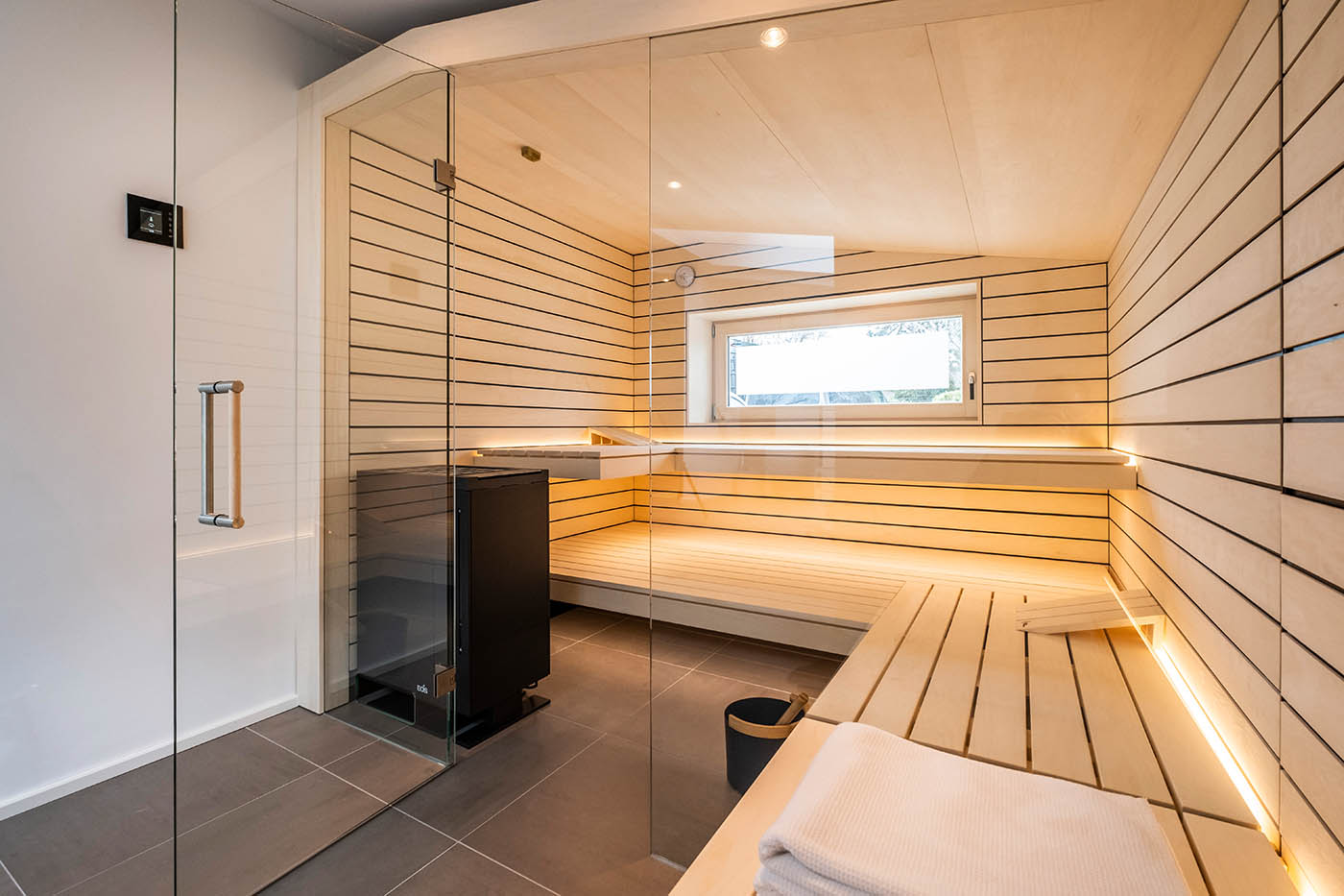 Sauna mit Fenster nach draußen 26 Design Sauna mit Fenster nach draußen. Sauna mit Dachschräge nach Maß in hellem Holz. Große Glasfront, nach außen verlängerte Bänke und LED Beleuchtung. Frei stehender Saunaofen und schwebende Saunabänke.