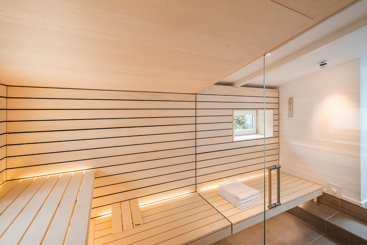 Sauna mit Fenster nach draußen 6 Design Sauna mit Fenster nach draußen. Sauna mit Dachschräge nach Maß in hellem Holz. Große Glasfront, nach außen verlängerte Bänke und LED Beleuchtung. Frei stehender Saunaofen und schwebende Saunabänke.