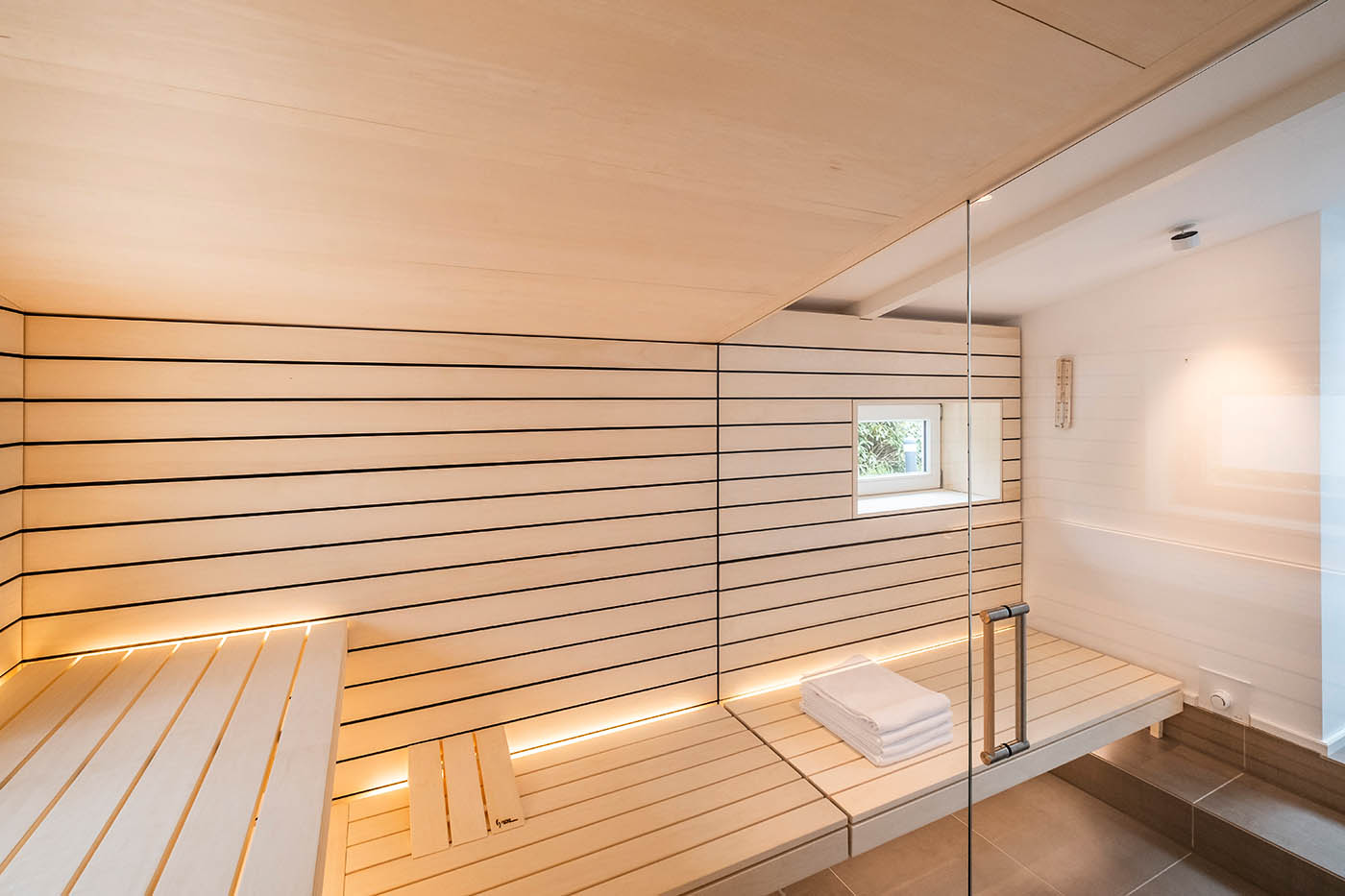 Sauna mit Fenster nach draußen 22 Design Sauna mit Fenster nach draußen. Sauna mit Dachschräge nach Maß in hellem Holz. Große Glasfront, nach außen verlängerte Bänke und LED Beleuchtung. Frei stehender Saunaofen und schwebende Saunabänke.